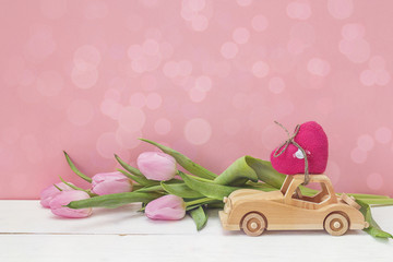 Wooden toy car carrying a heart and pink tulips on pink backgrou