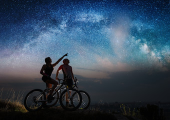 Fototapeta na wymiar Sporting pair with mountain bikes on the hill under night starry sky. Woman shows man at the stars. Night landscape with colorful Milky Way. Long exposure