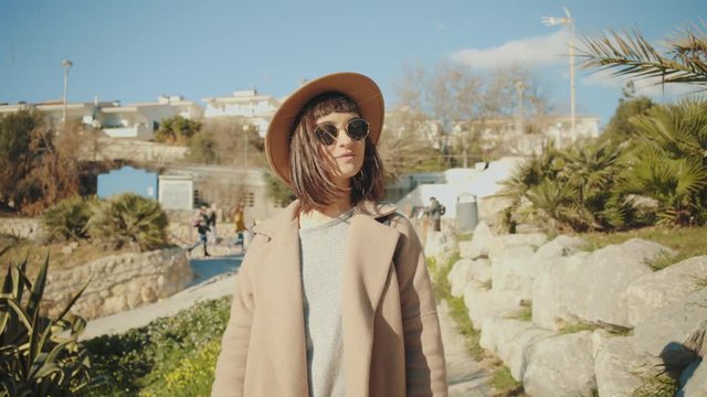 Camera follows in front attractive smiling brunette woman in sunglasses and wool trench walking down to sand beach next to sea through small narrow lane with green palms at sunny day.