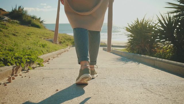 Camera follows behind girl legs in sneakers walking down on small narrow path with green grass on sides at sunny day.
