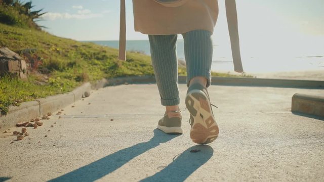 Camera follows behind girl legs in sneakers walking down and turns through small narrow lane with green grass on sides at sunny day.