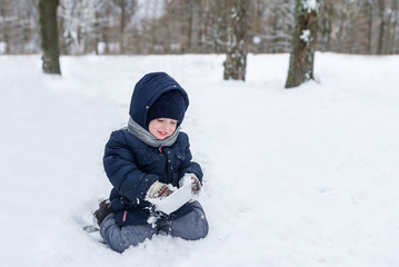 Fototapeta na wymiar Cute little boy sits in a snow in park in the winter. Boy cleans a plate snow.