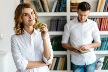Young man and woman in white shirts standing in a library. Woman holding an apple and looking at the camera. Behind the man reading a book. Many shelves with books on a blurred background
