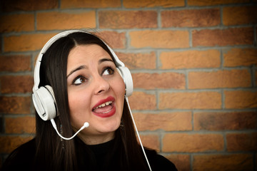 Happy beautiful young woman with headphones listening music and singing in front of the brick wall