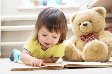 little girl reading book lying on stomach in her room on carpet with toy Teddy bear, smiling cute child, children education and development, happy childhood