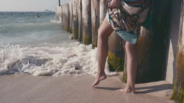 Close-up of women's legs go into the sea. Beautiful female ankle in the waves. Girl in dress comes to water (sea or ocean) and touch it by toes. Woman feet and legs walking on beach at sunset.