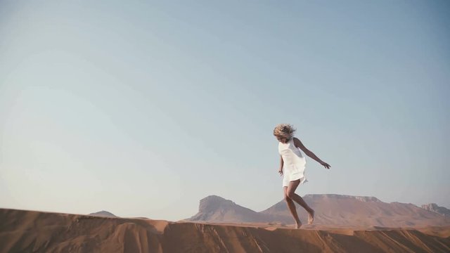 A young woman with blond hair - in the desert. Young woman in white dress jumping in the desert. She spread her arms, dress fluttering in the wind. Enjoying the wind. The feeling of freedom. Hot sand.