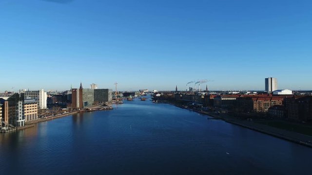A flight down Copenhagen harbor canal. Filmed on a calm and sunny day. Church and old building towers of Copenhagen sene in background. Filmed with a professional grade 4K drone.