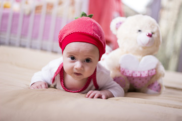 Close up of a beautiful baby girl lying on the bed. Infant lying on a bed. Family morning at home. Happy baby wearing strawberry shirt and pants. Newborn kid with toy bear.