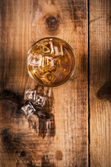 Glass of whiskey on wooden table with ice cubes around, view from above