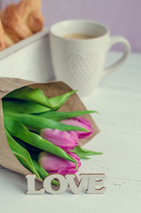 Cup of coffee with croissants, bouquet of pink tulips and wooden word LOVE