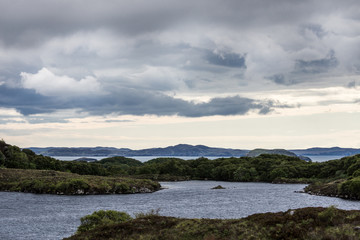 Northwest Coast, Scotland - June 6, 2012: Wide landscape of Loch A Chaim Bhaim irregular coastline under gray clouds. Green rocky rock formations in water upfront. Horizon of mountains.