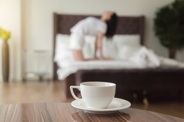 White cup with tea or coffee on table, woman practicing yoga at home on bed after waking up, working out, healthy start of a day, good early morning in hotel bedroom, refreshing before work concept