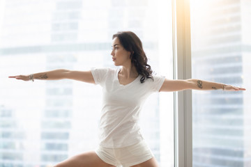 Young attractive woman practicing yoga in the morning, standing in Warrior Two exercise,...