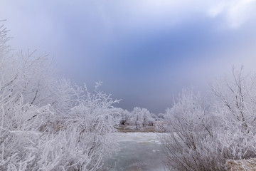 Beautiful winter scenery with trees covered by frost, along frozen river, and storm clouds