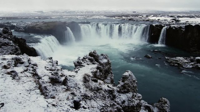 Godafoss waterfall in Iceland during a snowy, overcast morning shows the misty, eerie scenery as the water spills over the ledge.