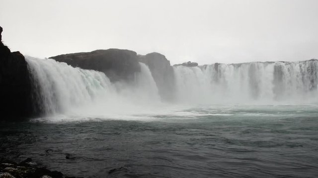 Godafoss waterfall in Iceland during a rainy, overcast morning shows the misty, eerie scenery as the water spills over the ledge.
