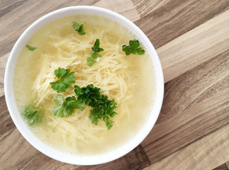 Chicken broth with noodles and green parsley. Meat chicken bouillon in a white bowl on wooden table. Top view.