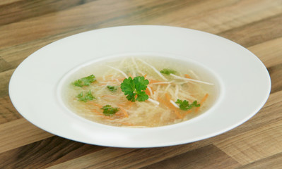 Chicken broth with carrot, celery and parsley. Rich chicken bouillon in a white plate on wooden table. Closeup view.