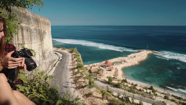 Young caucaasian woman taking pictures sitting on edge of a Melasti beach cliff overlooking roads and turquoise ocean on a sunny summer day in Bali, Indonesia. Shot with sony a7s on a slider