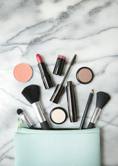 Aerial view of make up products spilling out of a pastel blue cosmetics bag, on a white marble counter top background