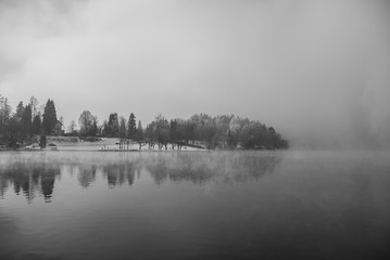 Fototapeta premium Island surrounded by fog reflecting in the water during the winter, lake Bled, Slovenia