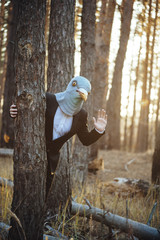 Creepy man in a suit and a rubber bird mask, standing in the sunset forest, behind the tree, waving...