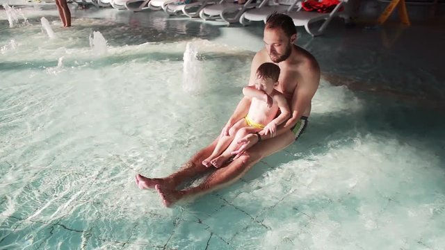 Young father and son in aquapark. Man with his cute baby boy sitting in a swimming pool and enjoying the waves