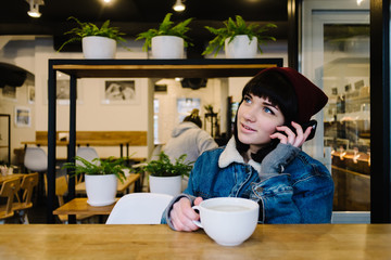 Young happy hipster girl talking on the phone and drinking a hot drink in cafe