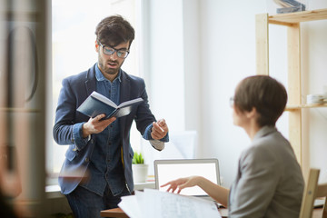 Portrait of contemporary young manager standing against window explaining work tasks to female colleague reading notes from planner book