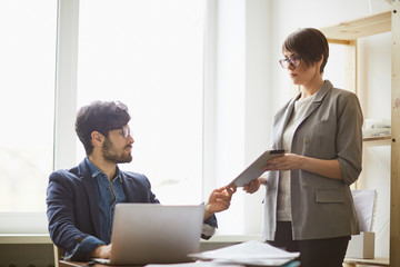 Fototapeta premium Young boss at workplace: successful middle eastern man looking up from laptop to check schedule at tablet in hands of his female colleague standing by him