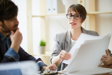 Portrait of business expert wearing creative haircut and glasses showing paperwork and explaining ideas to partner listening intently in modern office