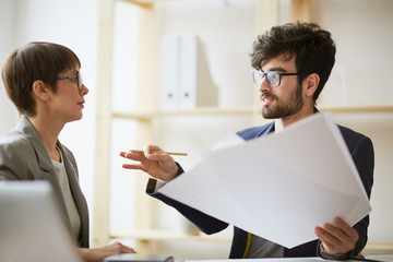 Portrait of bearded creative man holding heap of paper gesturing actively while explaining project details to businesswoman sitting next to him in modern office