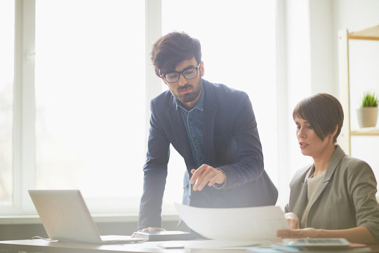 Successful young businesswoman showing plans to colleague leaning at her desk and discussing project