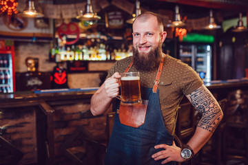 Bearded barman with tattoos wearing an apron standing near the bar and holding a glass of beer