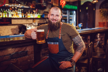 Bearded barman with tattoos wearing an apron sitting near the bar and holding a glass of beer