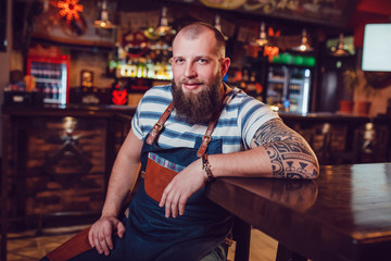 Obraz premium Bearded barman with tattoos wearing an apron sitting at the bar.