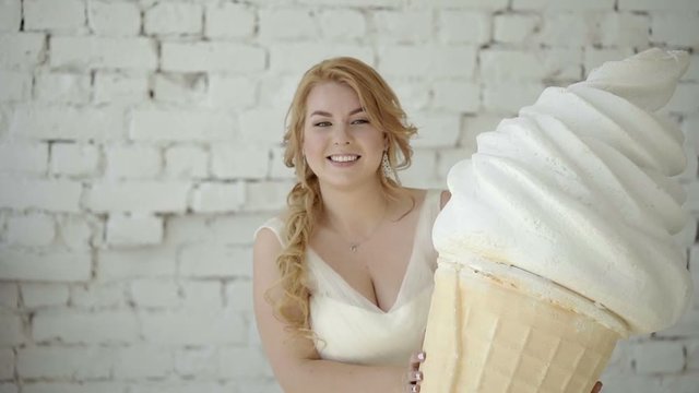 Cute young blonde woman in wedding dress posing in studio with huge ice cream shot