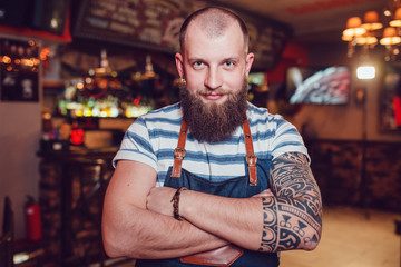 Bearded barman with tattoos wearing an apron standing at the bar with her hands folded.