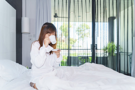 Young woman woke up and drinking coffee or tea on bed under sunlight