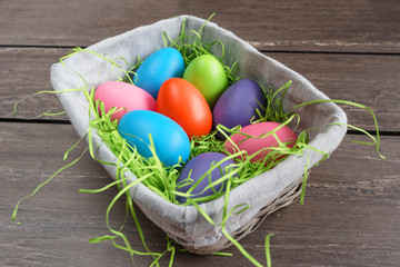 Easter wicker basket with colored eggs on grey wooden board.