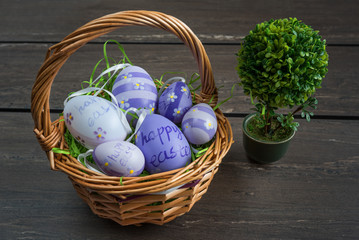 Easter wicker basket with colored eggs and a small bonsai on grey wooden board.