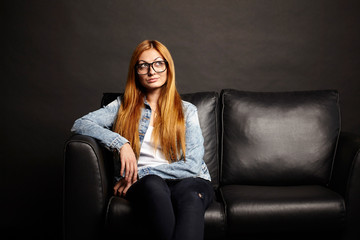 Obraz premium Attractive young woman in glasses sitting on black sofa on black background