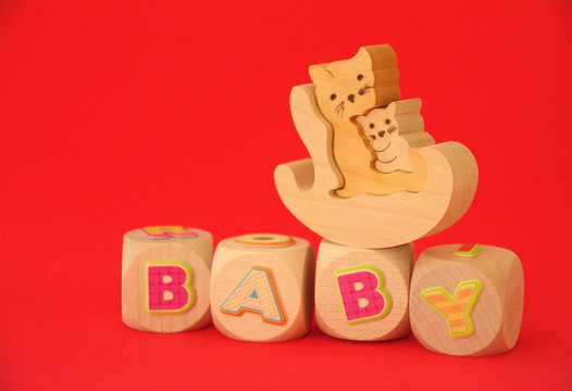 Wort Baby auf Holzw&uuml;rfeln mit Katze und Katzenbaby aus Holz im Schauckelstuhl auf rotem Hintergrund