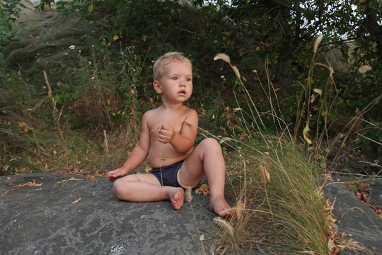 Small white Toddler sitting naked on a big gray stone and looks
