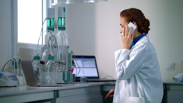 Woman scientist talking on phone in laboratory. Scientist in lab with modern laboratory equipment. Young researcher speaking on phone in chemical lab. Scientist woman talking on mobile phone