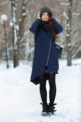 Beautiful winter portrait of young adorable redhead woman in cute knitted hat winter having fun on snowy park path