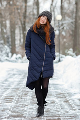 Beautiful winter portrait of young adorable redhead woman in cute knitted hat winter having fun on snowy park path