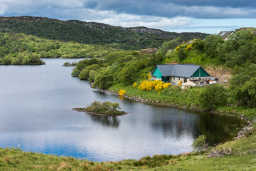 Fototapeta na wymiar Assynt Peninsula, Scotland - June 7, 2012: Cottage under construction at charming small lake with multiple green islands at Drumbeg hamlet. Blue sky, green hills, blue water, green pasture.