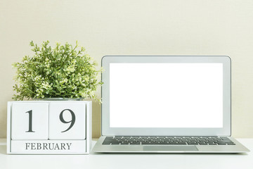 White wooden calendar with black 19 february word with white blank space at center of computer notebook on white wood desk and cream wallpaper textured background in selective focus at the calendar